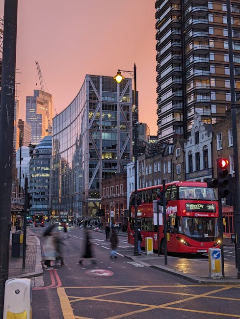 An urban street scene in Stratford, London, during dusk with a mix of modern glass office buildings and older brick structures. A red double-decker bus displaying 'Stratford City' on its destination sign is parked at a bus stop on the right side of the street. Several pedestrians, some blurred to indicate movement, walk across the crosswalks and along the pavement. On the left, a white and yellow pedestrian refuge island separates the road lanes, with traffic lights visible including a red light for vehicles. The street is equipped with street lamps, some lit, and road markings such as yellow box junctions and double-red lines indicating parking restrictions. Behind the bus, there are various vehicles and construction cranes visible against a pinkish evening sky. The scene illustrates a busy city environment suitable for house removals and moving services, with visible furniture, boxes, and packing materials possibly being loaded or unloaded nearby, although not directly shown in this shot, in an area prepared for home relocation activities. The image emphasizes transportation and urban moving logistics, aligning with the services provided by Man With a Van Stratford.