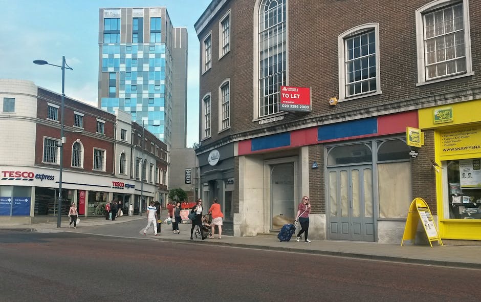 A street scene on Stratford High Street E15 showing a brick building with large arched windows and a blue, red, and gray striped storefront awning, which appears to be temporarily closed with a 'To Let' sign posted. To the right, there is a yellow shop with a stand-up sign outside and a window displaying various items. Pedestrians are walking along the sidewalk, some carrying shopping bags or rolling suitcases, with one woman in a pink top pulling a blue wheeled suitcase towards a doorway. Further left, a Tesco Express supermarket with large windows and the Tesco logo is visible, with people walking past. Behind, there's a modern high-rise building with a glass facade, contrasting with the older architecture. The street is well-lit with natural daylight, and a lamppost is seen on the pavement. The overall scene suggests a busy urban area with scheduled house removals or moving activities possibly occurring nearby, with focus on everyday transportation and pedestrian movement in Stratford.