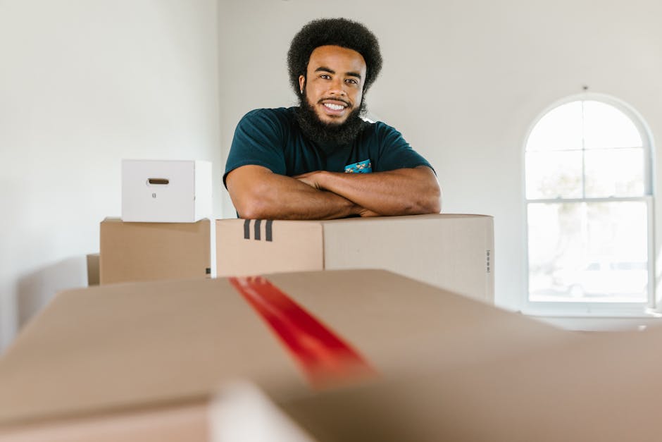A young man with dark skin, a full beard, and curly black hair smiling confidently while leaning on top of several cardboard boxes filled with belongings, positioned inside a bright, spacious room wit