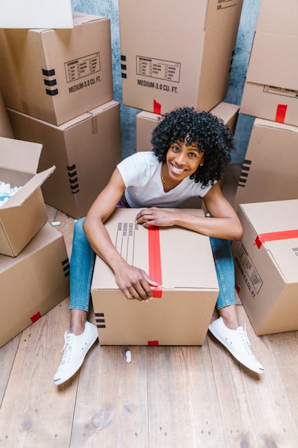 A young man with dark skin, a full beard, and curly black hair smiling confidently while leaning on top of several cardboard boxes filled with belongings, positioned inside a bright, spacious room wit