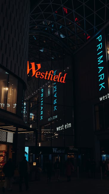 The image depicts the interior of Westfield Stratford City shopping centre in Stratford, with a high, curved glass ceiling and multiple illuminated signs displaying the Westfield and Primark logos in red and blue. The area is dimly lit, with silhouettes of people walking and standing near the entrances and shopping stores, including signs for West Elm. The scene suggests a busy retail environment, possibly during evening hours, with the architecture emphasizing modern design. This setting aligns with home relocation and packing activities managed by Man With a Van Stratford, highlighting the logistics of moving and furniture transport through a bustling commercial backdrop.